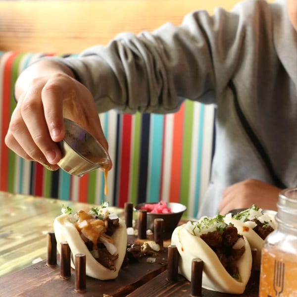 A person pouring sauce over a set of bao tacos on a wooden serving board.