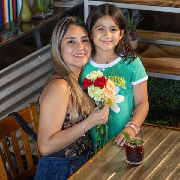 A woman and young girl smiling at the camera, holding flowers, with a drink on the table.
