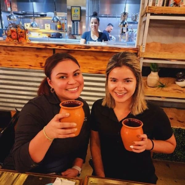 Two smiling women holding orange mugs, with a kitchen and another person in the background.