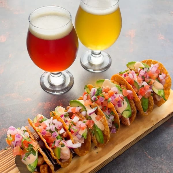 A wooden board holds seven tacos with avocado, pico de gallo, and sauce, next to two beers.