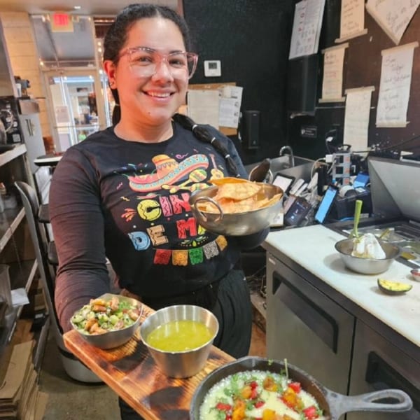 Smiling woman in a "Cinco de Mayo" shirt holding tortilla chips and a tray of taco appetizers.