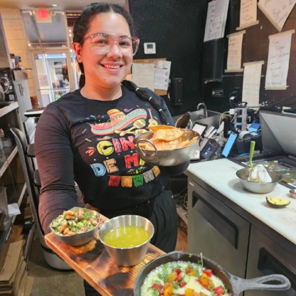 Smiling woman in a "Cinco de Mayo" shirt holding tortilla chips and a tray of taco appetizers.