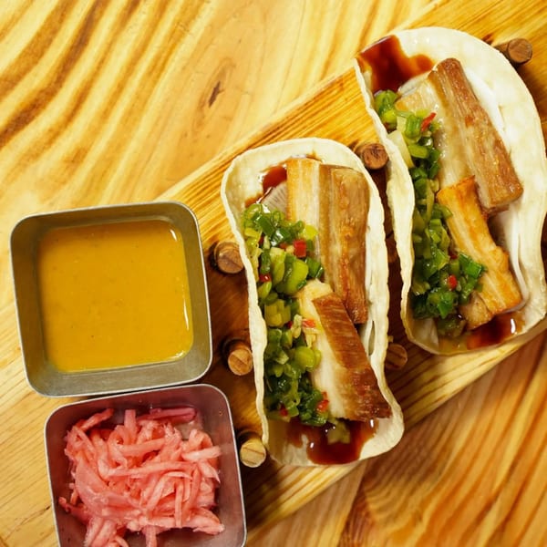 Two pork belly tacos with green relish and sauce, alongside pickled ginger and a yellow sauce.