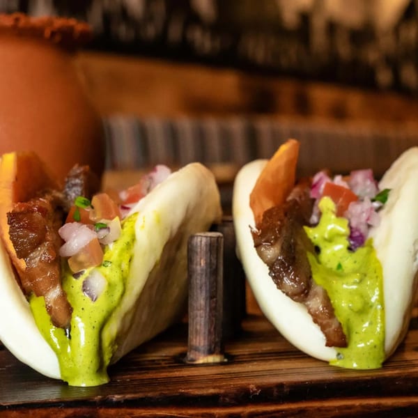Two bao bun tacos with grilled meat, pico de gallo, and green sauce on a wooden board.