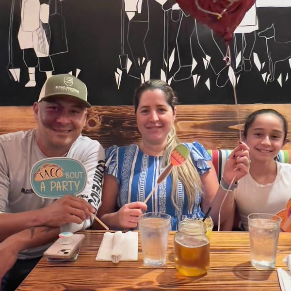 Family of three smiling, holding party props at a wooden table in A Taco by Divino.