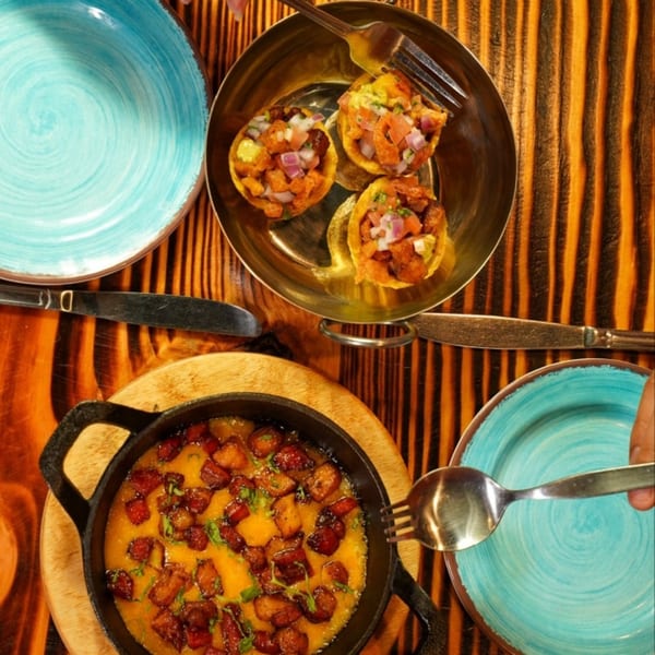 Overhead view of three mini tacos and a skillet of melted cheese with pork belly on a wooden table.