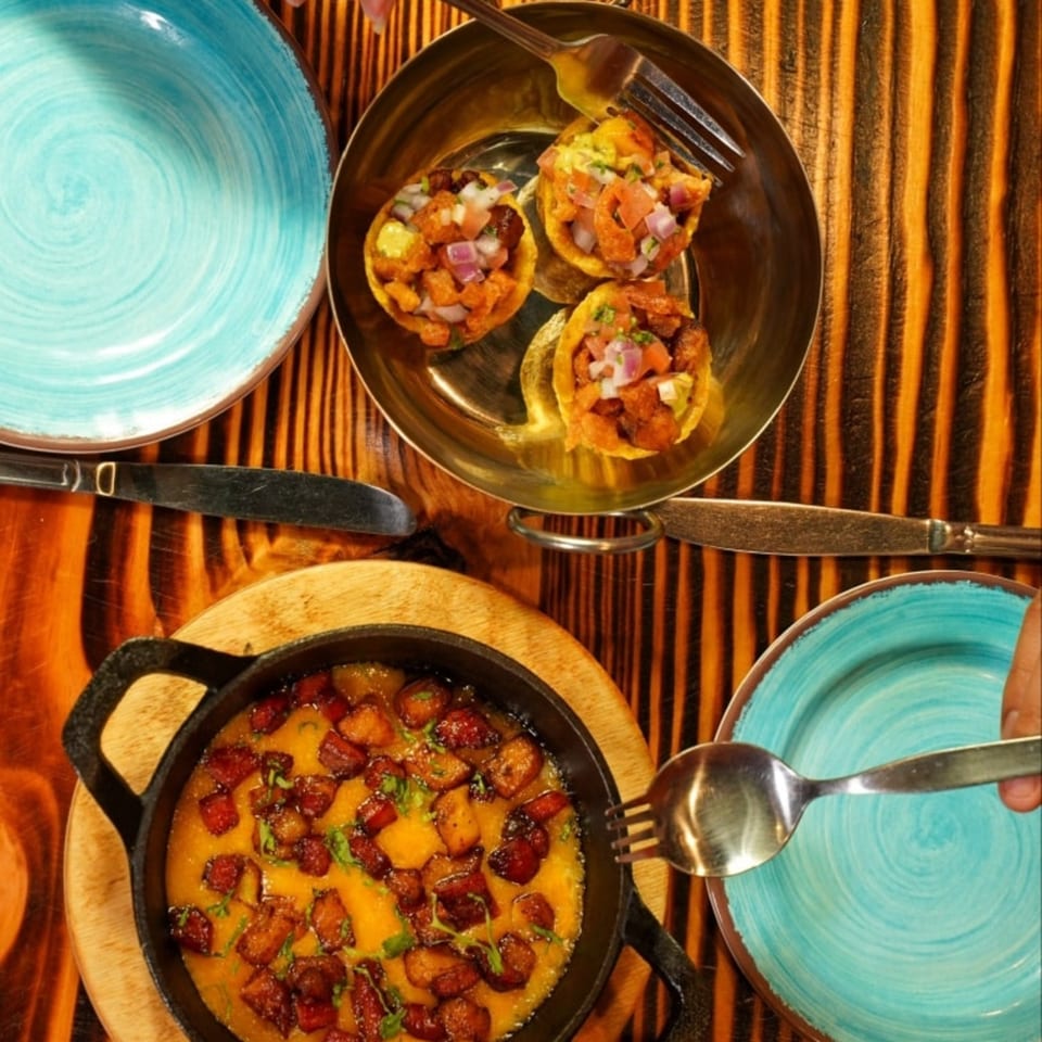 Overhead view of three mini tacos and a skillet of melted cheese with pork belly on a wooden table.